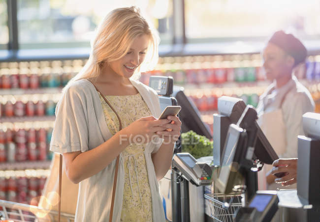 Sonriendo joven mujer mensajes de texto con el teléfono celular en la compra del mercado de comestibles - foto de stock