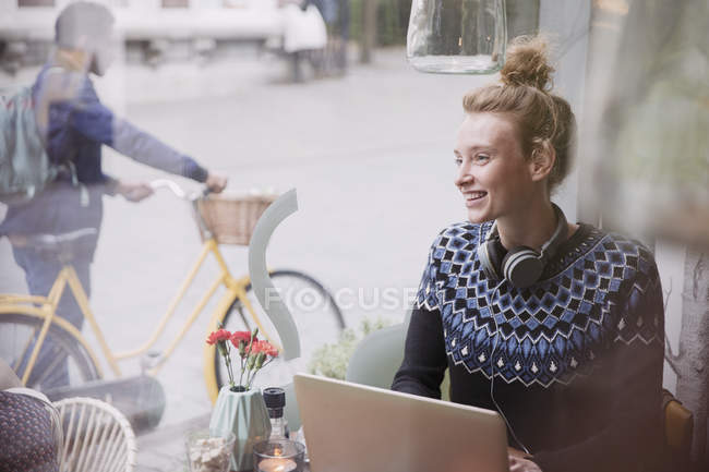 Lächelnde junge Frau mit Kopfhörern und Laptop im Fenster eines städtischen Cafés — Stockfoto