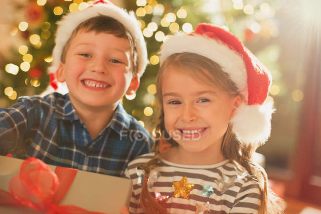Portrait smiling, enthusiastic brother and sister wearing Santa hats holding Christmas gift — Stock Photo