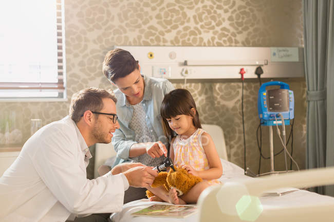 Male doctor using otoscope on teddy bear of girl patient in hospital room — Stock Photo