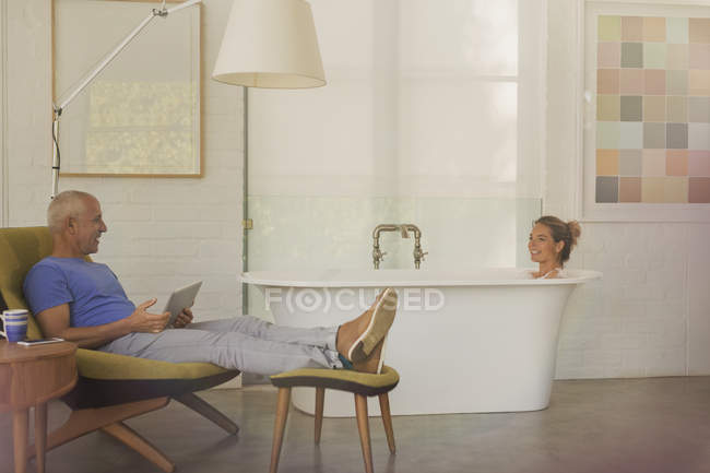 Husband with digital tablet relaxing, talking to wife in soaking tub in luxury hotel room — Stock Photo