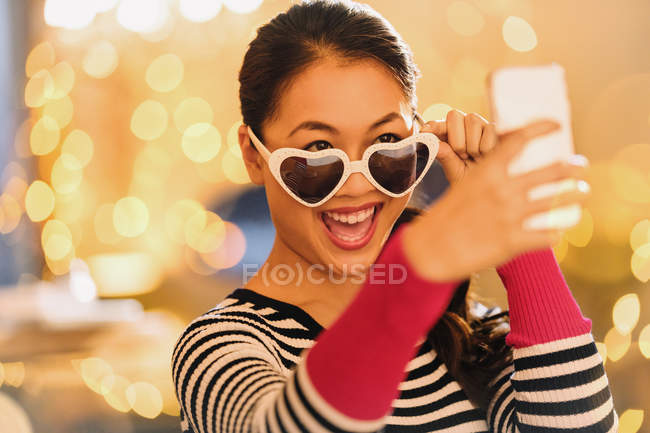 Mulher chinesa brincalhão usando óculos em forma de coração levando selfie com telefone da câmera — Fotografia de Stock