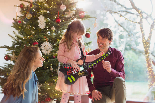 Родители смотрят, как дочь играет на игрушечной гитаре рождественский подарок перед елкой — стоковое фото