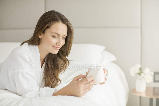 Femme souriante en peignoir buvant du café et envoyant des SMS sur son téléphone portable au lit — Photo de stock