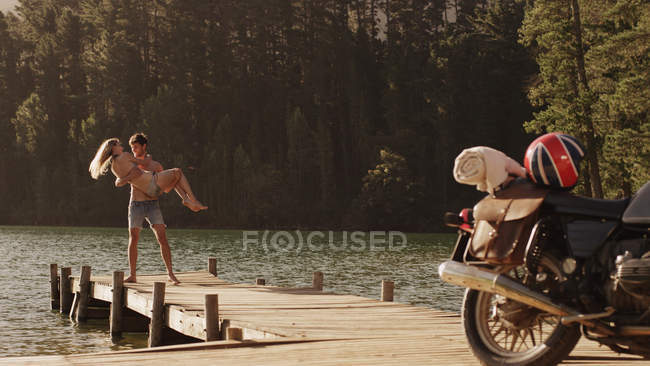 Jeune homme portant jeune femme sur quai au bord du lac près de la moto — Photo de stock