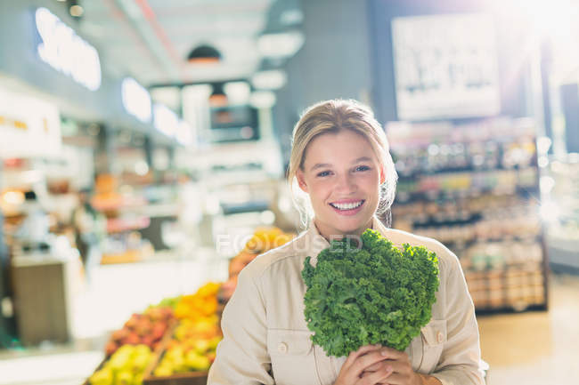 Retrato sonriente mujer joven sosteniendo racimo de col rizada en el mercado de comestibles - foto de stock