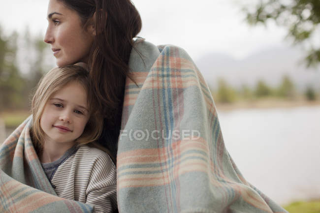 Портрет усміхненої дочки, загорнутої в ковдру з матір'ю на березі озера — стокове фото