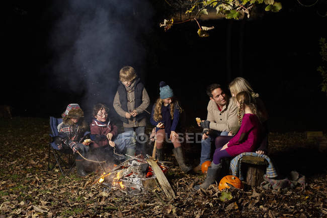Familia comiendo alrededor de la fogata por la noche en el bosque - foto de stock