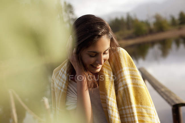 Mulher sorridente envolta em cobertor à beira do lago — Fotografia de Stock