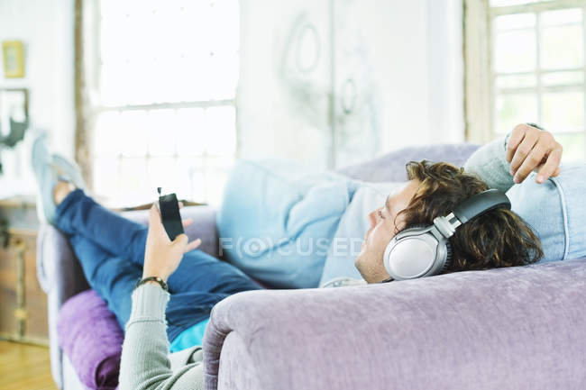 Joven escuchando los auriculares en el sofá - foto de stock