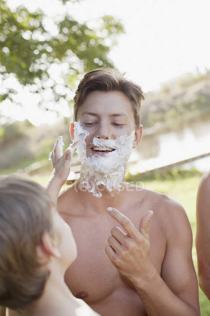 Сын помогает отцу наносить крем для бритья лицом к лицу на берегу озера — стоковое фото