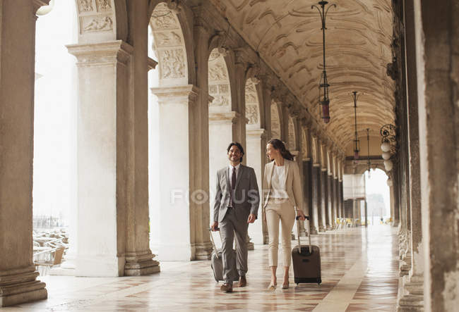 Hombre de negocios sonriente y mujer de negocios tirando de maletas a lo largo del corredor en Venecia - foto de stock