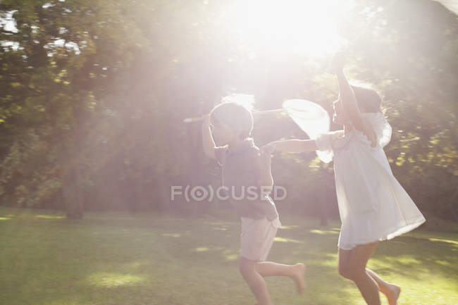 Niño y niña tomados de la mano y corriendo con redes de mariposa en la hierba - foto de stock