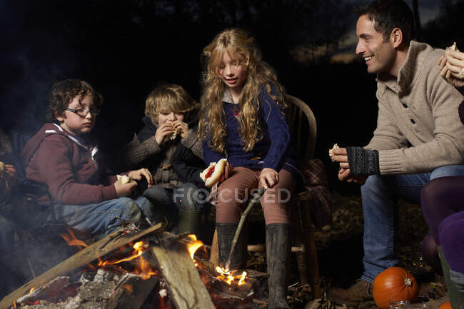 Familia comiendo alrededor de la fogata por la noche - foto de stock