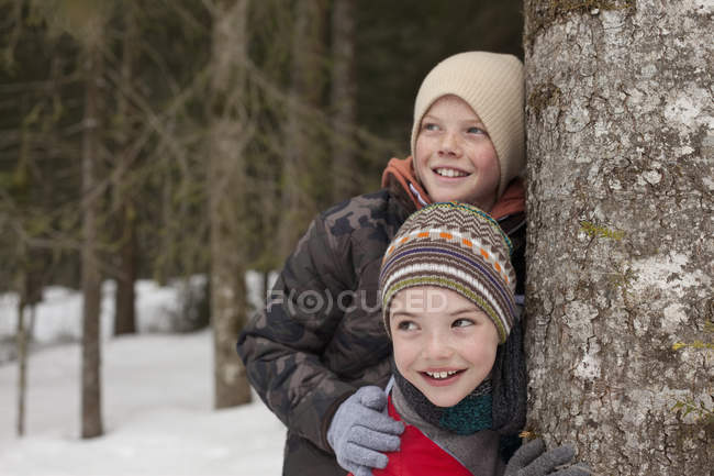 Meninos felizes encostados contra tronco de árvore em bosques nevados — Fotografia de Stock