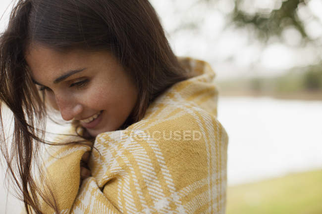 Primer plano de mujer sonriente envuelta en manta a orillas del lago - foto de stock