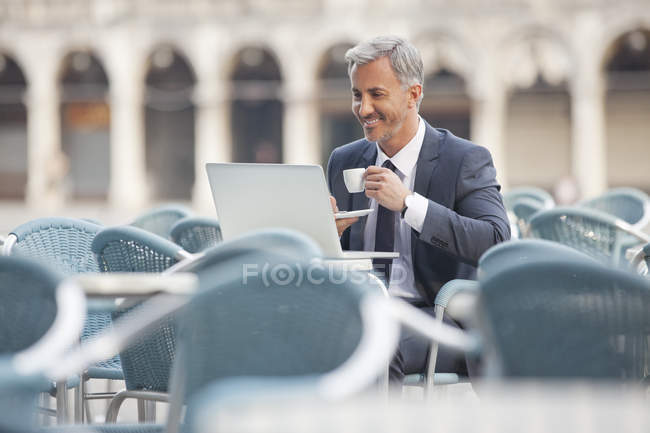 Hombre de negocios sonriente bebiendo café y utilizando el ordenador portátil en la cafetería de la acera - foto de stock