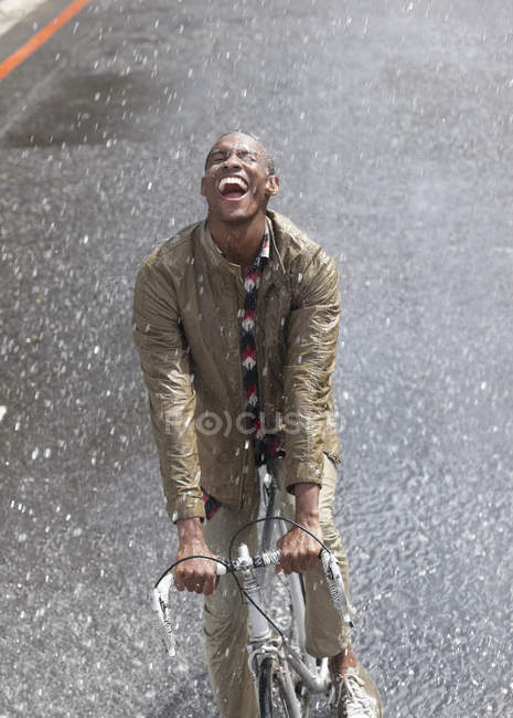Entusiástico homem negro andando de bicicleta na chuva — Fotografia de Stock
