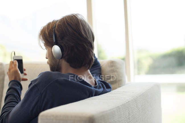 Hombre escuchando auriculares en el sofá, vista trasera - foto de stock