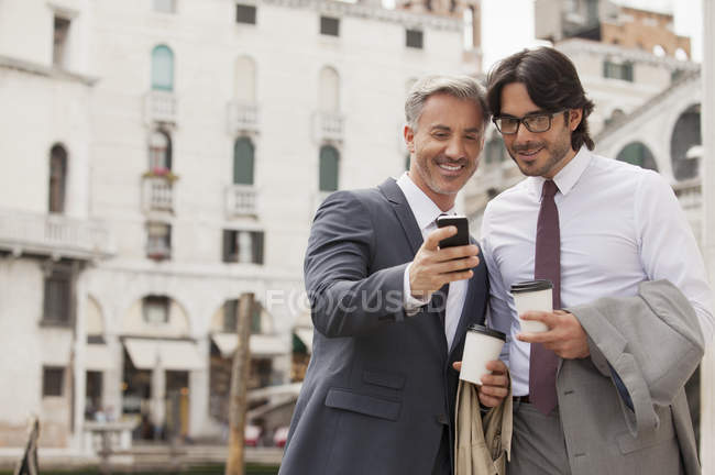 Hombres de negocios sonrientes con café mirando hacia abajo al teléfono celular en Venecia - foto de stock