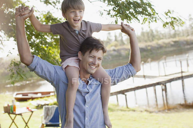 Отец носит улыбающегося сына на плечах у озера — стоковое фото