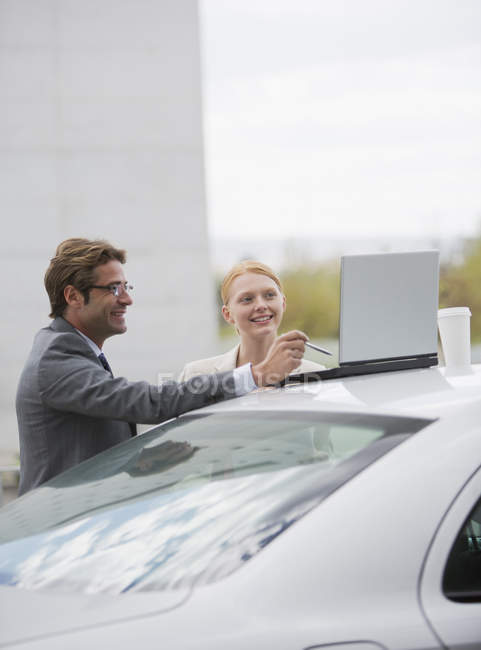 Hombre de negocios sonriente y mujer de negocios usando el ordenador portátil en la parte superior del coche - foto de stock