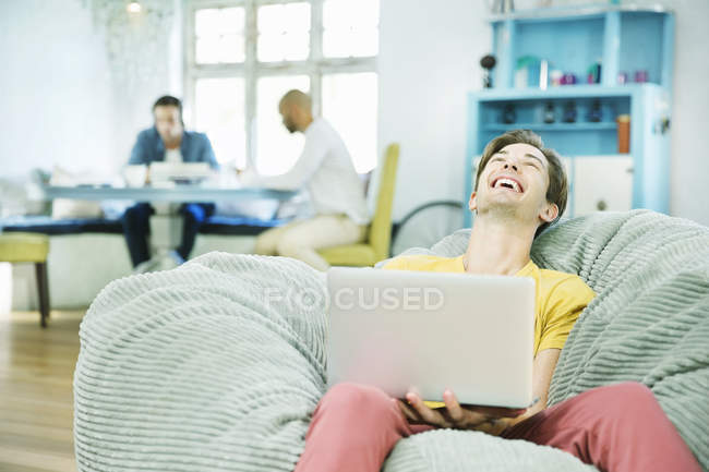 Riendo hombre usando el ordenador portátil en la silla del bolso de frijoles - foto de stock
