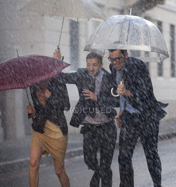 Gente de negocios feliz con sombrillas corriendo en la calle lluviosa - foto de stock