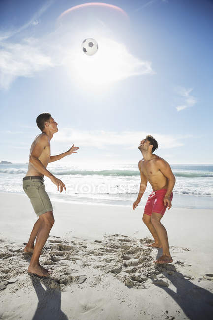 Homens em troncos de natação dirigindo bola de futebol na praia — Fotografia de Stock