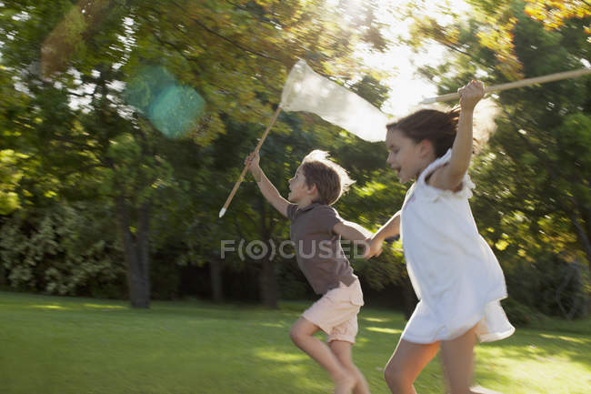 Happy boy and girl holding hands and running with butterfly nets in grass — Stock Photo
