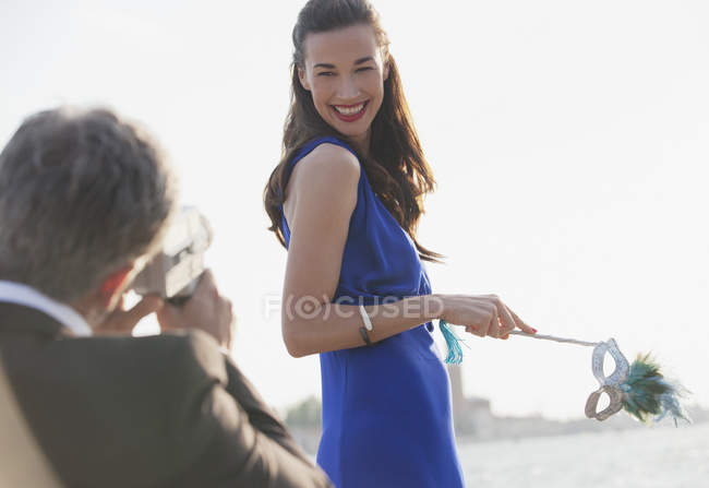 Hombre filmando sonriente mujer sosteniendo máscara en el paseo marítimo - foto de stock