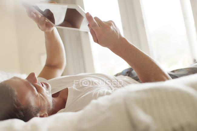 Усміхнений чоловік лежить на ліжку і використовує цифровий планшет — стокове фото