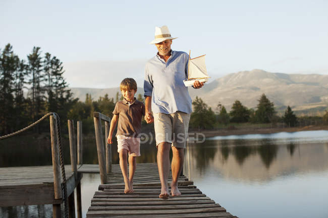 Улыбающиеся дедушка и внук с игрушечным парусником, держащимся за руки и идущие вдоль причала через озеро — стоковое фото