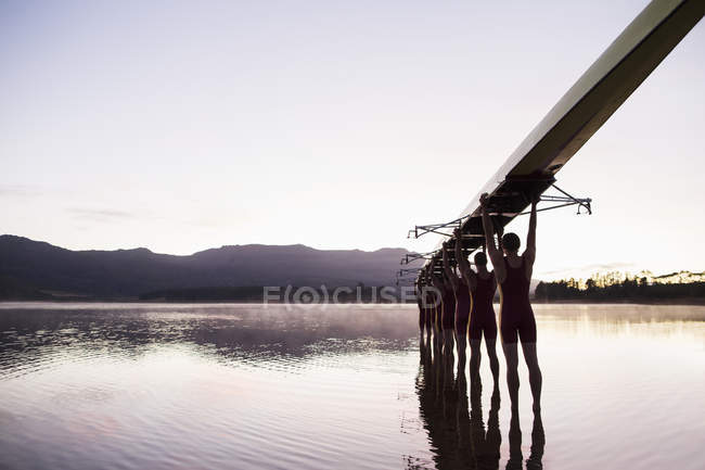 Equipo de remo entrando en el lago al amanecer con scull por encima - foto de stock