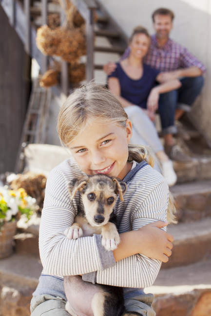 Retrato de menina sorridente segurando filhote de cachorro com os pais no fundo — Fotografia de Stock