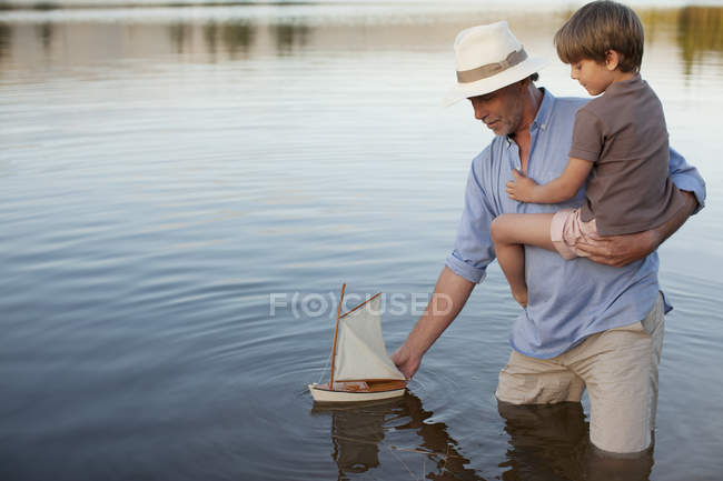 Grandfather and grandson wading in lake with toy sailboat — Stock Photo