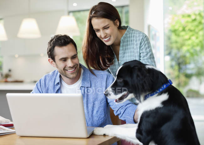 Pareja acariciando perro en la mesa con el ordenador portátil en casa moderna - foto de stock
