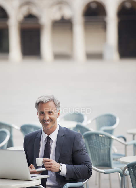 Retrato de homem de negócios sorrindo bebendo café expresso e usando laptop no café da calçada — Fotografia de Stock