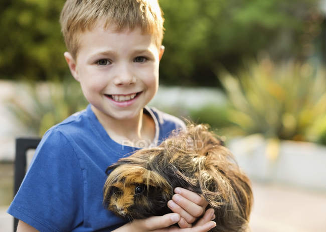 Smiling boy holding guinea pig outdoors, blurred background — Stock Photo