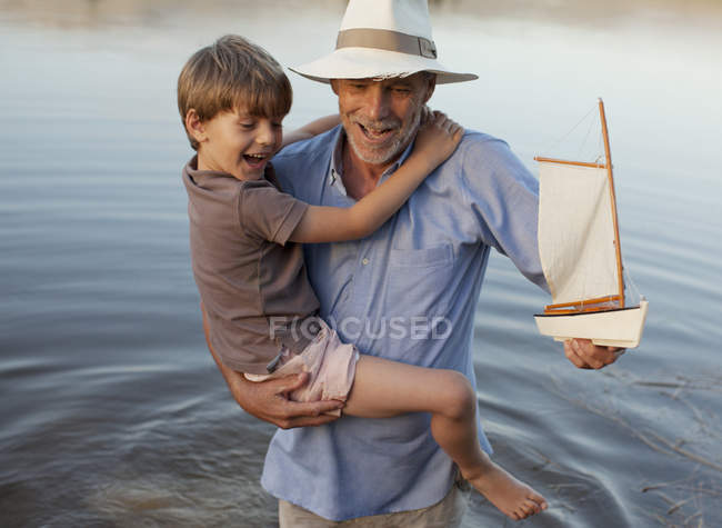 Grand-père et petit-fils souriants avec voilier jouet pataugeant dans le lac — Photo de stock