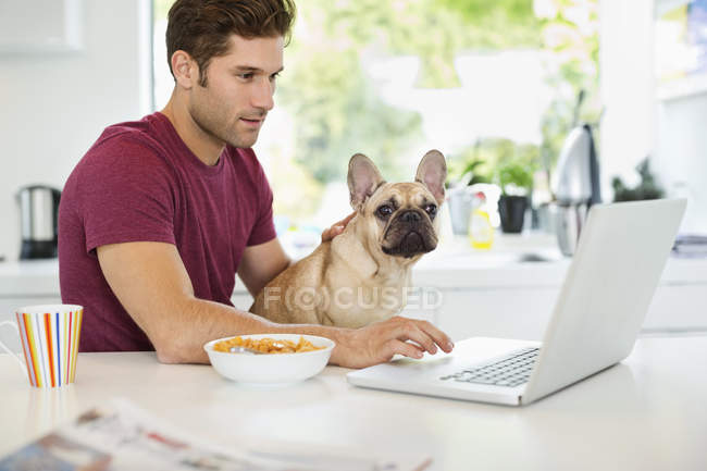 Человек на ноутбуке ласкает собаку на кухне в современном доме — стоковое фото