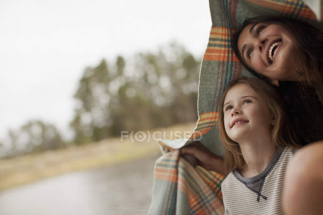 Madre e hija sonrientes bajo una manta a orillas del lago - foto de stock