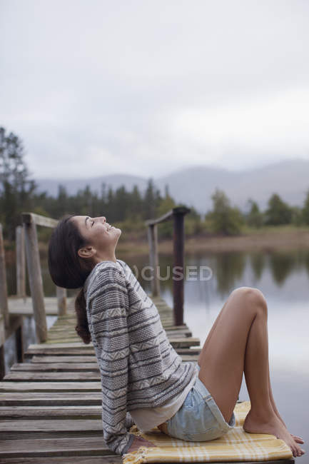 Mulher sorridente sentada na doca à beira do lago com a cabeça para trás — Fotografia de Stock