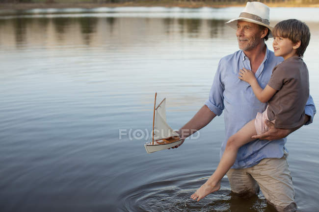 Grandfather and grandson with toy sailboat in lake — Stock Photo