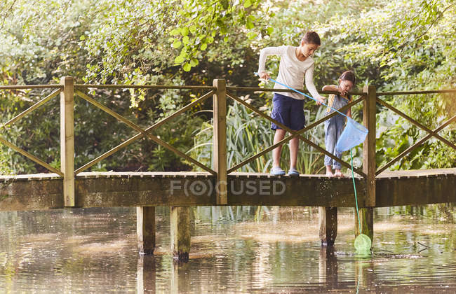 Brother and sister fishing with nets at footbridge over pond — Stock Photo