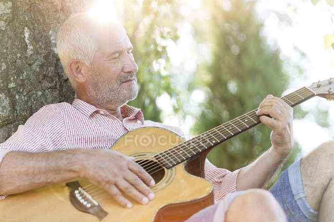 Senior man playing guitar against tree trunk — Stock Photo