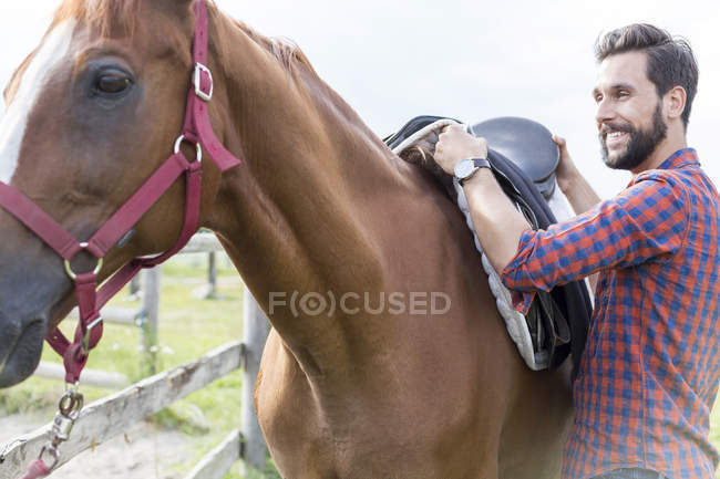 Улыбающийся человек снимает седло с лошади — стоковое фото