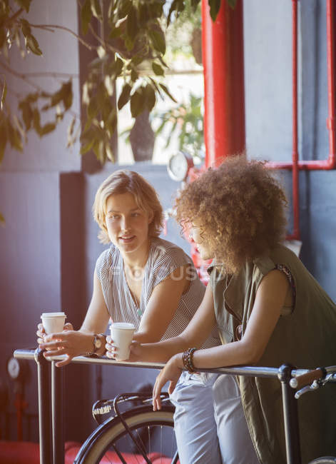 Mujeres bebiendo en el café apoyadas en barandilla en la tienda de bicicletas - foto de stock