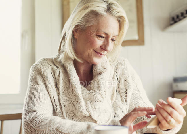 Улыбающаяся пожилая женщина в свитере переписывается с телефоном на кухне — стоковое фото