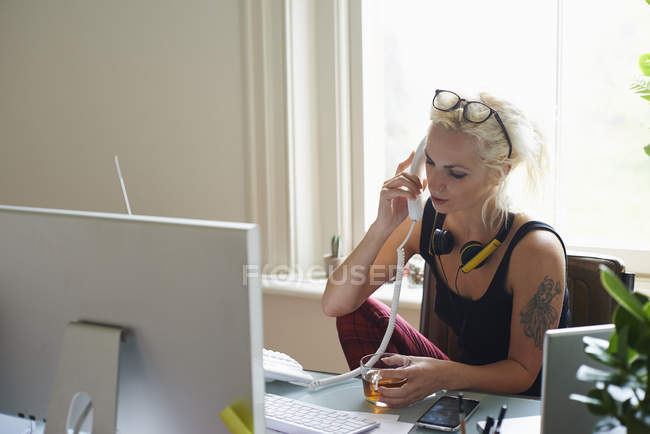 Молодая женщина с наушниками и татуировкой пьет чай и разговаривает по телефону в домашнем офисе — стоковое фото